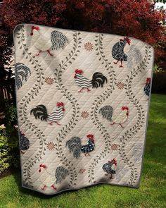 a quilt with roosters on it sitting in the grass next to some trees and bushes