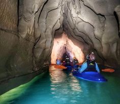 two people in kayaks are entering a cave with blue water and green rocks on either side