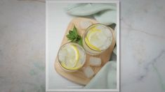 two glasses filled with ice and lemon on top of a cutting board next to a towel