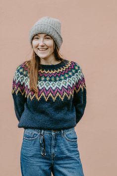 a smiling woman wearing a knitted sweater and jeans stands in front of a pink wall