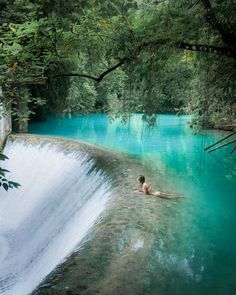 a person swimming in a river with blue water and green trees around it, surrounded by greenery