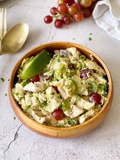 a wooden bowl filled with chicken salad next to silverware and grapes on a table