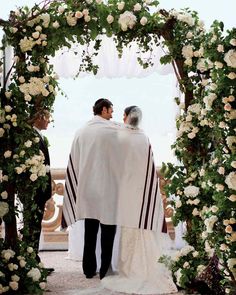 a man and woman standing in front of an archway with flowers on the sides, dressed in white