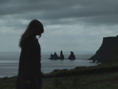 a woman standing on top of a lush green hillside next to the ocean under a cloudy sky