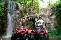 two people standing on four wheelers in front of a waterfall with an animal statue behind them