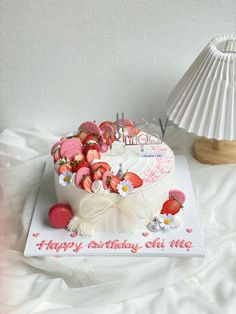 a white birthday cake with strawberries and flowers on it sitting on a table next to a lamp