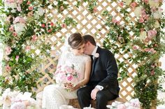 a bride and groom are sitting on a bench in front of a floral arch with roses