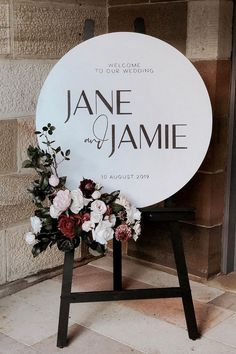 a wedding sign with flowers on it in front of a brick wall