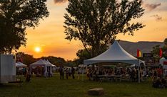 a group of people standing around tents in the middle of a grass field at sunset