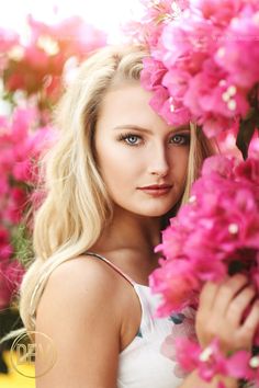 a beautiful blond woman with blue eyes and long hair standing in front of pink flowers