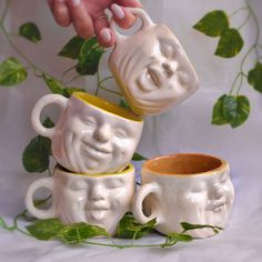 three coffee mugs with faces on them being held by someone's hand over some leaves