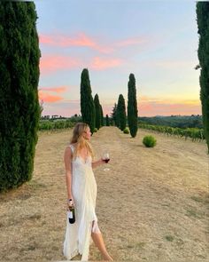 a woman in a white dress holding a glass of wine while standing on a dirt road