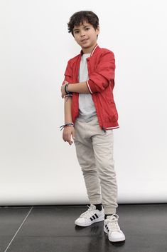 a young boy standing in front of a white wall wearing a red jacket and grey pants