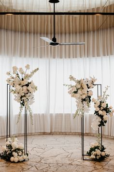 two tall vases with white flowers and greenery in front of a curtained window