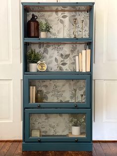 a blue bookcase with books and plants on it in front of a wallpapered background