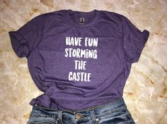 "A fun, and soft shirt for fans of The Princess Bride. This comfy shirt makes an awesome gift and goes well with everything from jeans and boots to leggings and sneakers. *Processing time is currently 4-9 business days. Thank you for your patience!* Design: \"Have Fun Storming The Castle\" Shirt Sizing Softstyle (Unisex Adult T-Shirt) - Comfortable for both women and men to wear. - Please see size chart in the photos for more details. Materials - 100% preshrunk ringspun cotton - Heather colors 3 Casual Purple T-shirt For Fan Merchandise, Casual Pre-shrunk Shirt For Fan Conventions, Casual Purple T-shirt For Fans, Fun Slogan T-shirt For Fan Merchandise, Casual Shirt With Funny Text For Fans, Casual Shirt With Letter Print For Fan Conventions, Casual Tops With Funny Text For Gifts, Casual Purple T-shirt As Gift, Casual Purple Slogan T-shirt