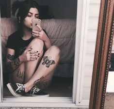 a woman with tattoos sitting on the floor in front of a mirror looking at her cell phone