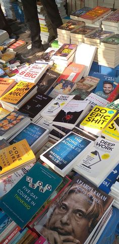 many books are stacked on top of each other with one person standing in the background