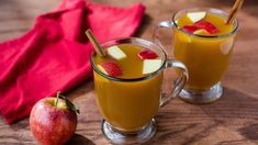 two mugs filled with orange juice and apples on a wooden table next to a red napkin