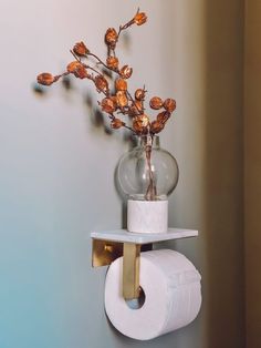 a toilet paper holder with flowers in a glass vase on the wall next to a roll of toilet paper