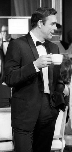 a man in a tuxedo is holding a coffee cup and looking at something