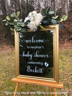 a welcome sign for a baby shower with flowers on the frame and greenery around it