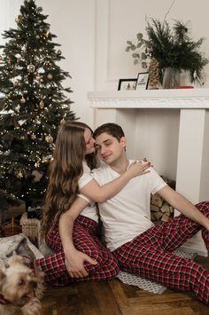 a man and woman sitting on the floor in front of a christmas tree with their dog