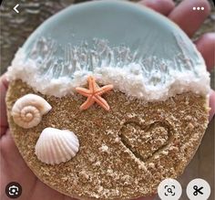 a hand holding a piece of sand with shells and starfish on it in the shape of a heart