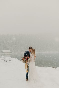 a bride and groom are standing in the snow near a lake on their wedding day