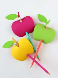 three apples and two pencils on a white surface with paper clips attached to them