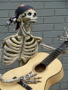 a skeleton holding a guitar and wearing a bandana on it's head, standing in front of a brick wall