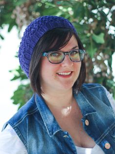 a woman wearing glasses and a blue knitted hat smiles at the camera while standing in front of a tree