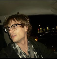 a man wearing glasses sitting in the back seat of a car at night with city lights behind him