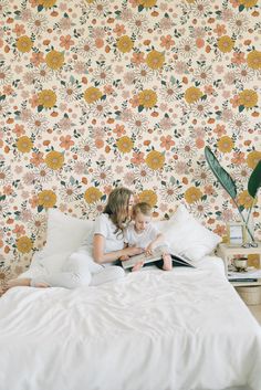 a mother and daughter sitting on a bed reading a book in front of a floral wallpaper