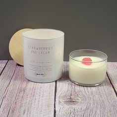 a white candle sitting next to a glass on top of a wooden table with a strawberries and cream label