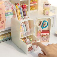 a hand is pointing at some stationery items in a holder on a desk next to other office supplies