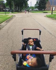 two small children sitting in a stroller on the side of a road with houses behind them