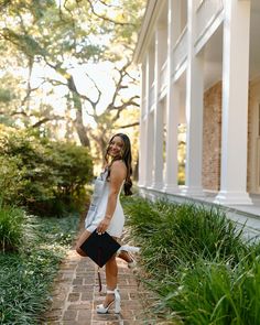 a woman walking down a brick path in front of a building with columns and trees