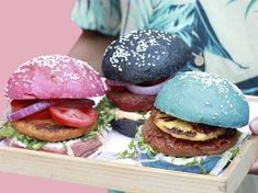 three burgers with different toppings are in a wooden tray on a pink background