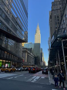 people crossing the street in front of tall buildings with skyscrapers on both sides and cars driving down the street