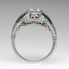 This superlative ring features a pierced design with milgrain details. It is centered with a .79 carat round brilliant cut diamond in a square bead setting. The top and shoulders of the ring are bead set with a total of forty-two (42) round old European and round brilliant cut diamonds. The shoulders are each channel set with six (6) calibré cut emeralds. The ring measures 15.4mm at the top, rises 7.5mm above the finger, and tapers to 1.8mm wide by 1.2mm thick at the base of the shank. It is currently a size 7.5. Art Deco Diamond Emerald Ring With Brilliant Cut, Art Deco Emerald Ring With Diamond, Art Deco Emerald Ring With Diamond Center Stone, Art Deco Emerald Diamond Ring With Accents, Art Deco Emerald Ring With Brilliant Round Cut, Art Deco Brilliant Cut Emerald Ring, Art Deco Emerald Ring With Round Cut In Platinum, Art Deco Emerald Ring With Round Cut Center Stone, Luxury Emerald Ring With Single Cut Diamonds For Anniversary