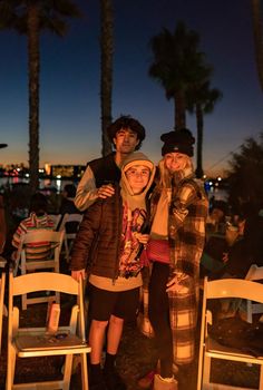 two people standing next to each other in front of some chairs and palm trees at night