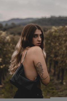 a woman with tattoos on her back standing in the middle of a field and looking off into the distance