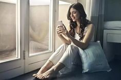 a woman sitting in front of a window looking at her cell phone while holding a pillow