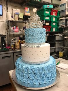 a three tiered cake with blue frosting and a tiara on top, sitting on a counter in a kitchen