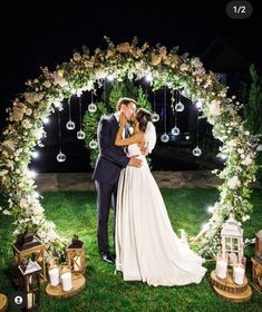 a bride and groom kissing in front of an archway decorated with white flowers at night