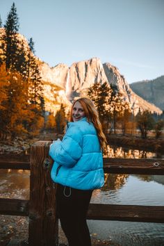 a woman in a blue puffy jacket leaning on a wooden fence near a lake