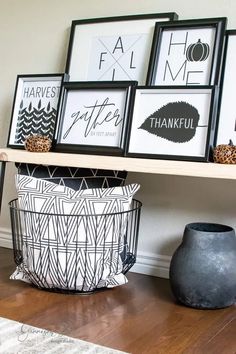 some framed pictures are on a shelf above a basket and a black vase with a pumpkin in it