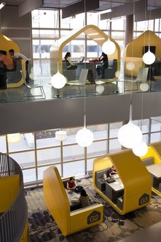 people are sitting at desks in an office with yellow and gray furniture, large windows, and hanging lights