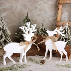 two white reindeer standing next to each other in front of snow covered trees and pine cones